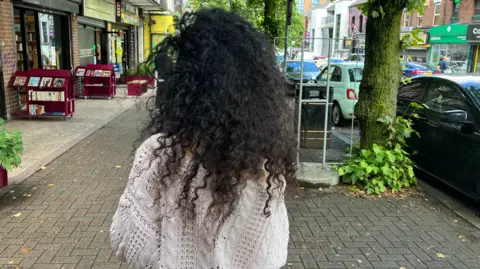 Woman with curly black hair with her back to the camera standing on a street with cars parked along the road