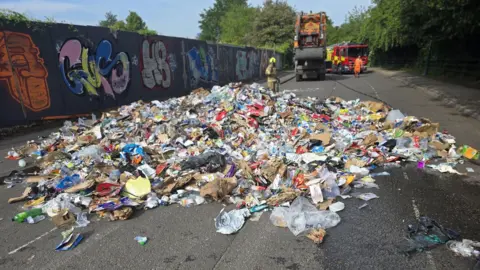 Nottingham City Council Six tonnes of rubbish has been emptied from a bin lorry all over a road after a fire took hold in the back of a bin lorry. You can see the collection vehicle in the background with a fire engine parked behind it. There is one firefighter standing behind the rubbish and a refuse collector in orange in front of the fire engine. 