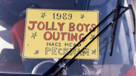Only Fools and Horses Appreciation Society A close-up of a handwritten sign in a coach windscreen reading “1989 Jolly Boys Outing, Nag’s Head Peckham”, with windscreen wipers across the glass.