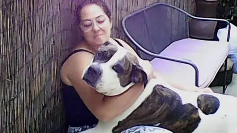 Eve Williamson Eve holds American Bulldog Khan in her arms while sitting on a chair in the garden.
