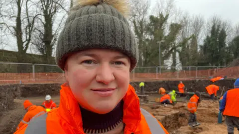 Penny Jackson wearing a hi-vis jacket and a green woollen hat. She is standing in the dig site. About 10 people are standing in the dig site working.