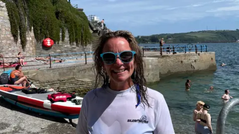 A woman wearing blue sunglasses and a white rash vest smiles at the camera. Her hair is wet. Behind her people are swimming in the sea.