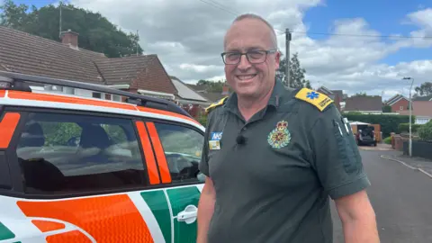 A man wearing a green ambulance uniform standing in front of a yellow and green branded community first responder car