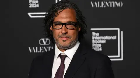 David Olusoga looking into the camera. He has dark, jaw-length hair and is wearing dark glasses and a suit. He is standing in front of a black background with the Valentino and International Booker Prize 2024 logos.