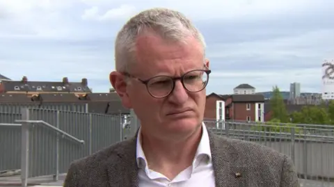 Pol Callaghan has short grey hair and black-rimmed glasses and is wearing a sports jacket and open-necked white shirt. A metal fence and houses are in the background.