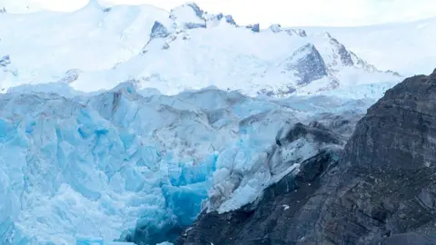 A large glacier next to a large rock, with a river basin below
