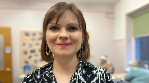 A women is smiling at the camera. She has short brown hair and is wearing a black and white dress. She has large hooped earrings made up of different sections in each ear