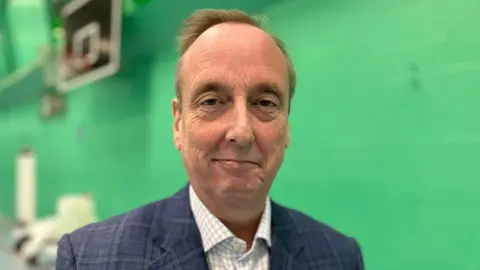 Ian Thorn smiles at the camera - a head and shoulders shot, he wears a blue jacket and checked shirt. Blurred behind him is a blurred green wall.