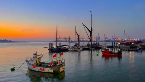 Walking Tractor/BBC Weather Watchers Small fishing boats are floating on the water, and industrial port buildings and cranes are on the horizon. The picture is taken at dusk, with a partially orange sky.