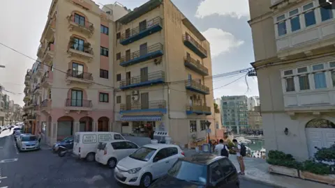 A road of hotels and tiled apartments overlooking the sea. Cars are parked outside. The bottom of one building has a blue and white striped awning and shop, and the other has pink arches and columns. 
