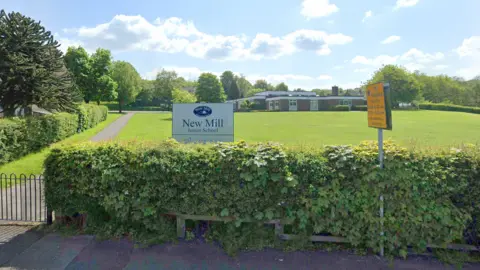 Google The entrance to New Mill Junior School in Holmfirth, with a sign displaying the school's name, and the school in the background across a grassy field.