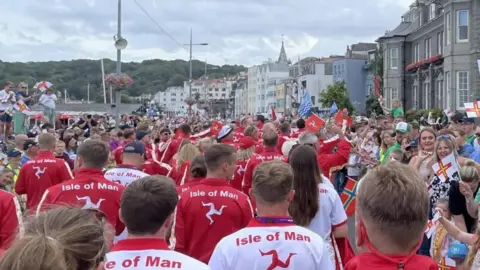 A sea of people in white and red Isle of Man tracksuits wave flags and wave at crowds lined along the seafront in Guernsey's St Peter Port.
