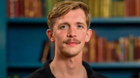 A young man with blonde hair and a moustache, pictured from the neck up, standing in front of a colourful bookcase