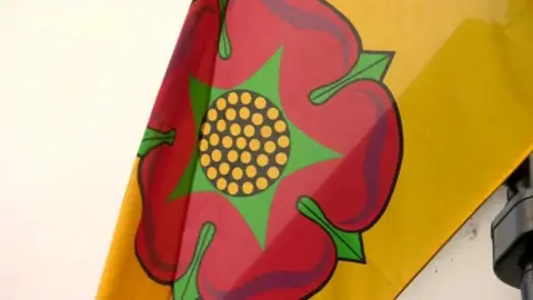 An image of a Lancashire flag flying. The flag is yellow with the Lancashire Red rose on it.
