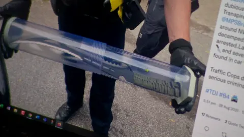 Metropolitan police A computer screen displaying a police social media post, including a photo of an officer holding a transparent tube (used to store evidence) containing a large knife with a curved blade and a serrated edge