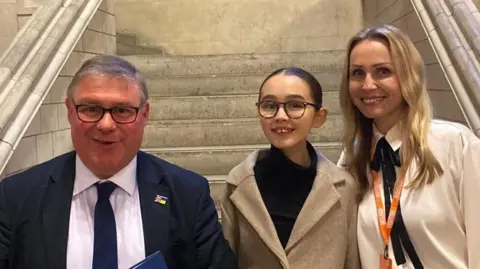 Mark Francois Mark Francois, Skye and Christina all standing in front of a white staircase in Parliament. Francois is wearing a navy suit jacket, white shirt and navy tie. Skye is wearing a tan jacket with a black top. Christina is wearing a white shirt. They are all smiling.