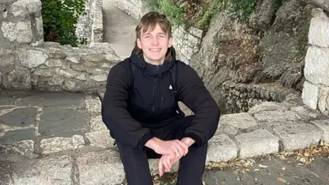 A young man with blonde hair smiles as he sits on a stone ledge at a ruined site. His hands are resting on his knees and he is wearing a black coat and black trousers