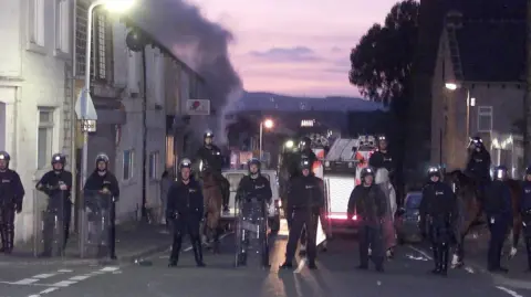 PA Media Police officers in full riot gear and shields block off a road by standing a in a line stretched across it. Smoke rises next to houses and a fire engine is behind police.