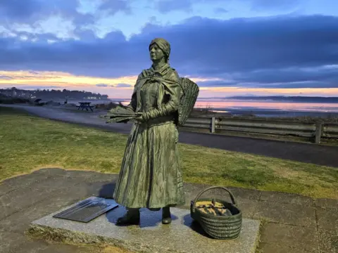 Moira MacKintosh A bronze statue of a late 19th Century fishwife holding fish in her hands and a creel on her back, with a bronze basket at her feet. The beach is in the background.