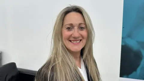 Ben Parker/BBC June Mummery with long hair smiling at the camera, she is wearing a white top and dark suit jacket.