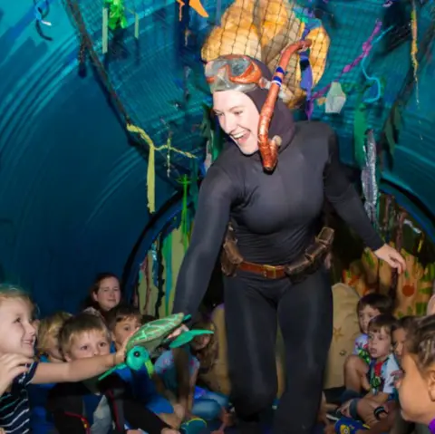 South Kesteven District Council A performer dressed as a deep-sea diver, wearing goggles and a snorkel. She is  with a group of children inside a decorated space with ocean-themed items. She holds a small sea turtle puppet. There are underwater decorations hanging from the blue, netted ceiling. 