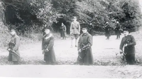 Getty Images/Bettmann A black and white archive photo of armed police guarding a rural border road in Northern Ireland circa 1922. Four officers in peaked caps and coats are standing in a shallow trench in the foreground, pointing guns. More officers are standing guard in the background. 