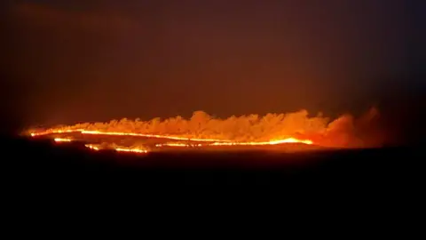 Elin Freeborn-Jones Orange wildfire can be seen ripping across the length of a hillside. The flames glow bright yellow and smoke can be seen rising from the line of fire. 