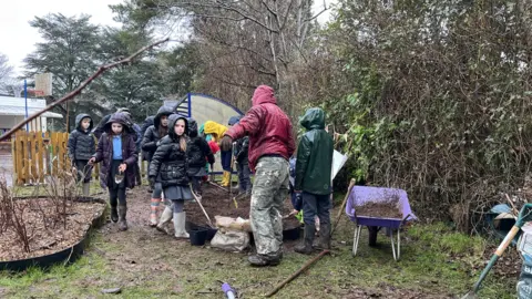 A group of children wearing rain coats and wellies. They are standing in a school garden on a rainy day, digging holes into a bed of soil. There are wheelbarrows full of soil to the right, and a shovel leaning on bags of dirt. 