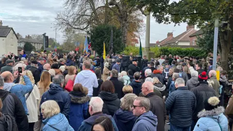 People in coats stand around in the cold to mark the event at the memorial at Brough