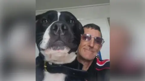 Suffolk Police A close-up photo of a black and white dog's head. Next to it is a man wearing glasses. Both are looking into the camera. 