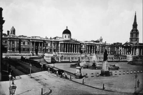 Getty Images A black and white image from 1886 showing Trafalgar Square with the National Gallery along the back of the square beside St Martin-in-the-Fields. Horse and carts are lined up along one road