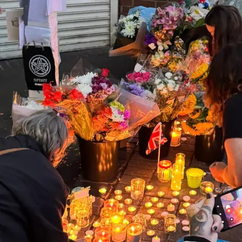 Three women kneel down next to a number of lit candles on the ground. Behind the candles are a number of bunches of flowers in varying colours.