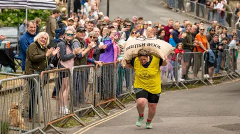 Aircam Drone Services @aircamdrone.co.uk A woman wearing a yellow top carries a bag of wool over her shoulders as she runs along a road as part of the Tetbury Woolsack Races. Dozens of people are watching her from alongside the road, behind metal barriers
