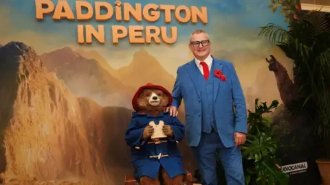 PA Media Hugh Bonneville poses for a photo with Paddington Bear in front of a large poster advertising the new film Paddington in Peru. He wears a light blue suit with matching waistcoat and trousers, a bright red tie and a red poppy on the lapel. He has thick dark glasses and is smiling. paddington is holding a sandwich with a chunk bitten out, and wears his blue duffel coat and red hat. The poster shows mountains and jungle in the background.