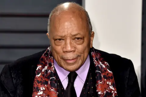 Getty Images News photo of Quincy Jones, smartly dressed in a dark suit, purple shirt and black tie, accessorised with a brown abstract-pattern silk scarf. He is quite elderly in this photo, with no hair and a thin moustache.