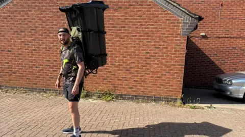 Daniel Fairbrother Daniel Fairbrother, standing by a red brick wall, with shorts and a T-shirt on, with a large black wheelie bin on his back. He is side-on. He is wearing a cap that is on the other way round.