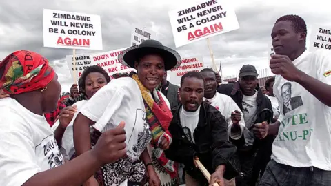 AFP/Getty Images A crowd Zanu-PF supporters dance together as they welcome President Robert Mugabe home from a trip abroad in 2000, with placards saying ‘Zimbabwe will never be a colony again’.