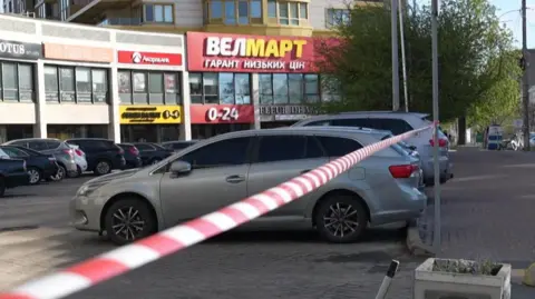 A mid shot of a block of shops with several cars parked in the car park. Police cordon tape is hung between two poles in the foreground. 