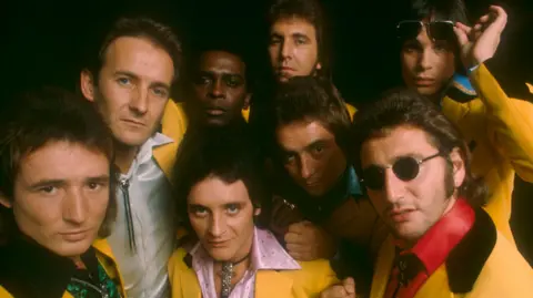 The members of Showaddywaddy dressed in yellow suits looking at the camera. 