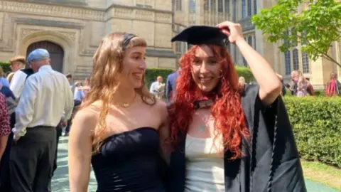 The two sisters are in the grounds of the Wills building on a sunny summer's day wearing sleeveless graduation dresses. One of the girls has dyed red hair and is wearing a graduation mortar board and gown