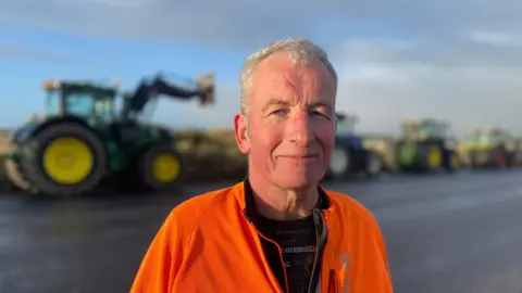 Farmer Duncan Wight wearing a bright orange jacket and black t shirt with text on it. He is stood in front of four tractors that have parked in the background,. He has short grey hair.