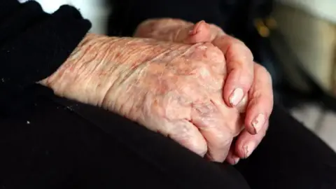 BBC An elderly woman with her hands placed on her lap, she is wearing black trousers. Her nails are painted in light pink nail varnish, which is chipped at the edges.