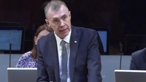 Senedd Cymru Adam Price, a man in a navy suit, speaks inside parliament.