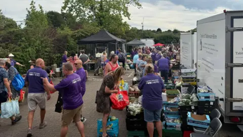 Towcester Community Larder A large group of people at a community food larder, with gazebos on the left, vans to the right, and boxes of food at the front. Helpers are wearing purple T-shirts. There are trees in the distance. 