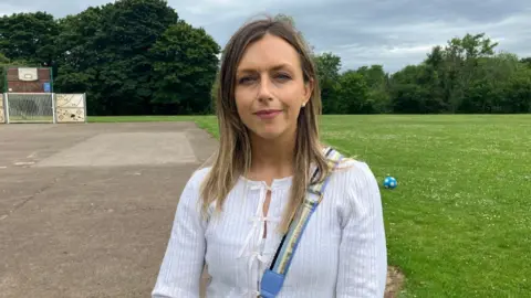 Laura has long dark hair and is wearing a long-sleeved white top with bows on the front, with a bag strap over her shoulder. She is standing in the park with the concrete surface behind and the grass beyond.