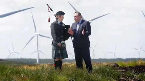 PA Media Former first minister of Scotland, Alex Salmond, is talking to a bagpiper. They are standing on grass in front of a large wind farm.