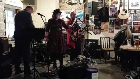 Kissing the Flint A band performs in a small pub with a woman in a dark dress and hat standing in front of a microphone between two guitarists