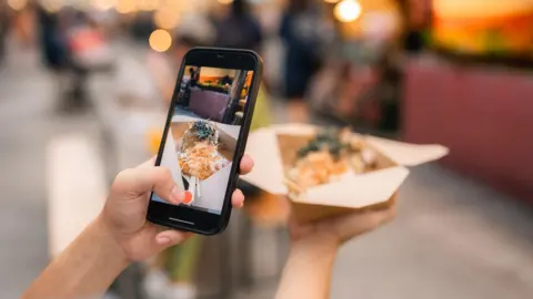 Getty Images A person is taking a photo of food they are holding with an iPhone. The food is in a brown cardboard container and has orange cheese on top and green herbs. There are market stalls blurred in the background