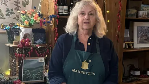 Mannetti's A woman wearing an apron stands behind a counter in a cafe. The apron reads "Mannetti's".