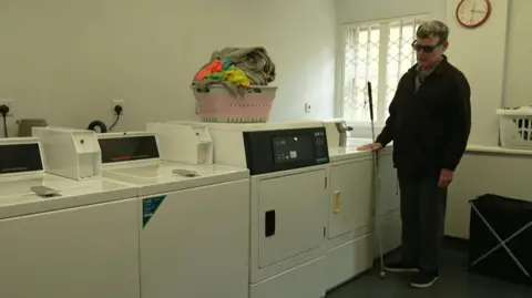 Andrew is standing beside four washer/dryers that have been adapted to be higher for blind people to use easily